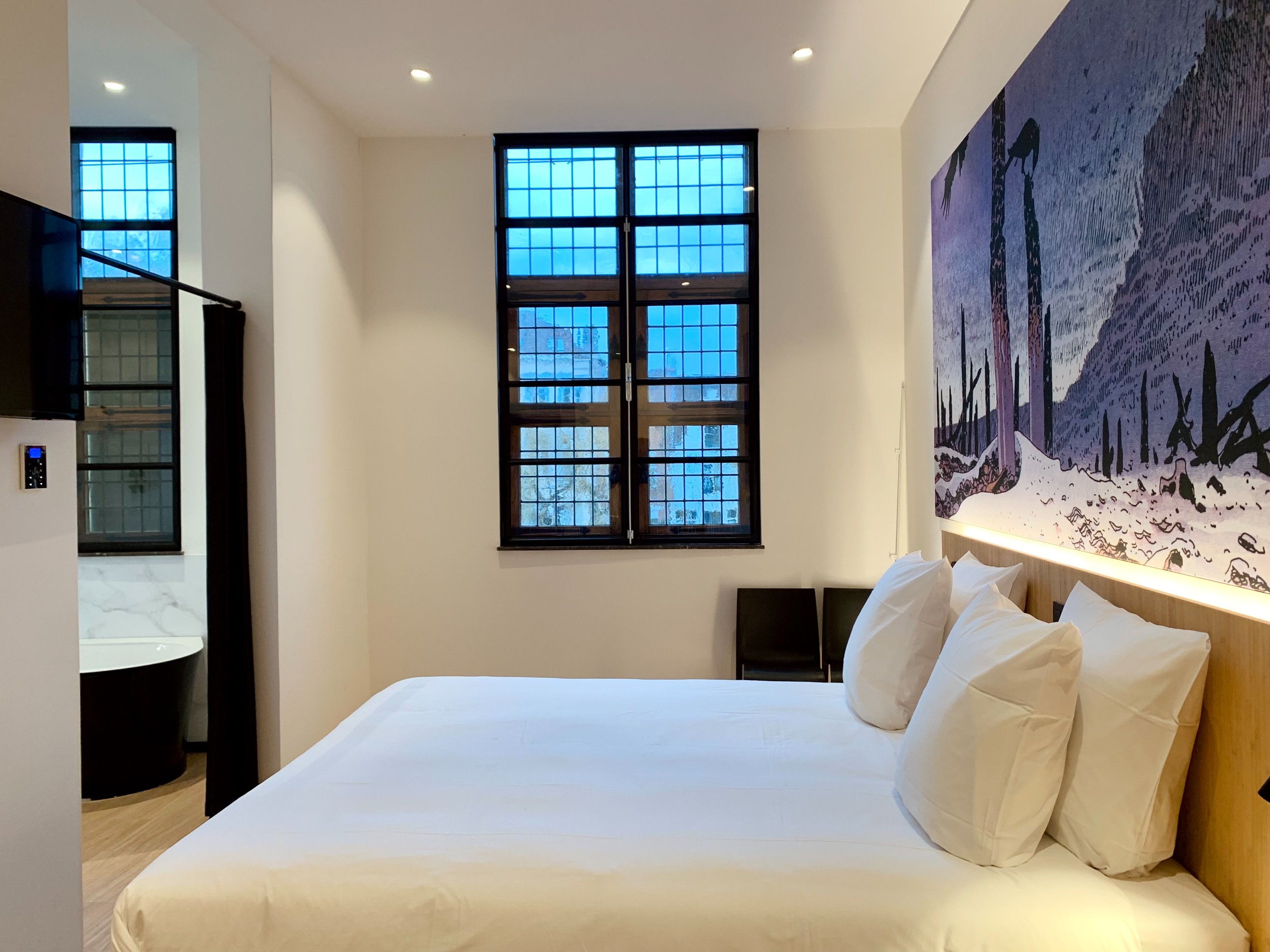 Modern hotel bedroom suite with contemporary artwork, large windows, and marble bathroom.